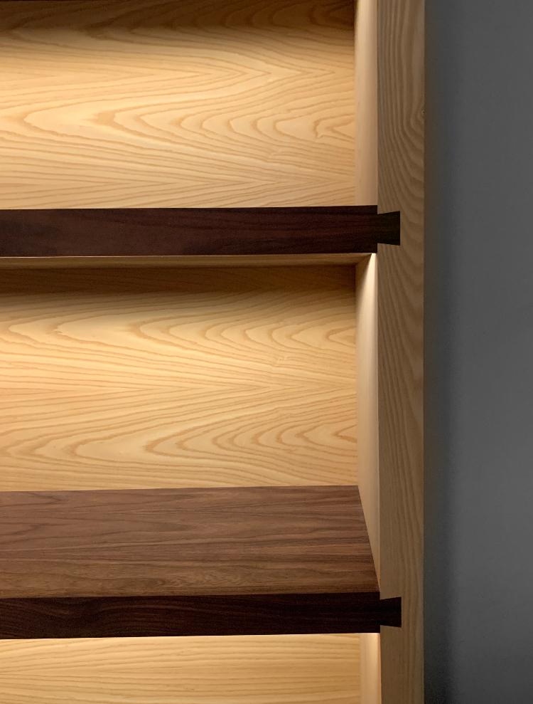 Close-up of a bespoke bookshelf with solid American walnut shelves joined by exposed dovetail joinery into a solid white oak case, with warm integrated LED lighting revealing the joint work