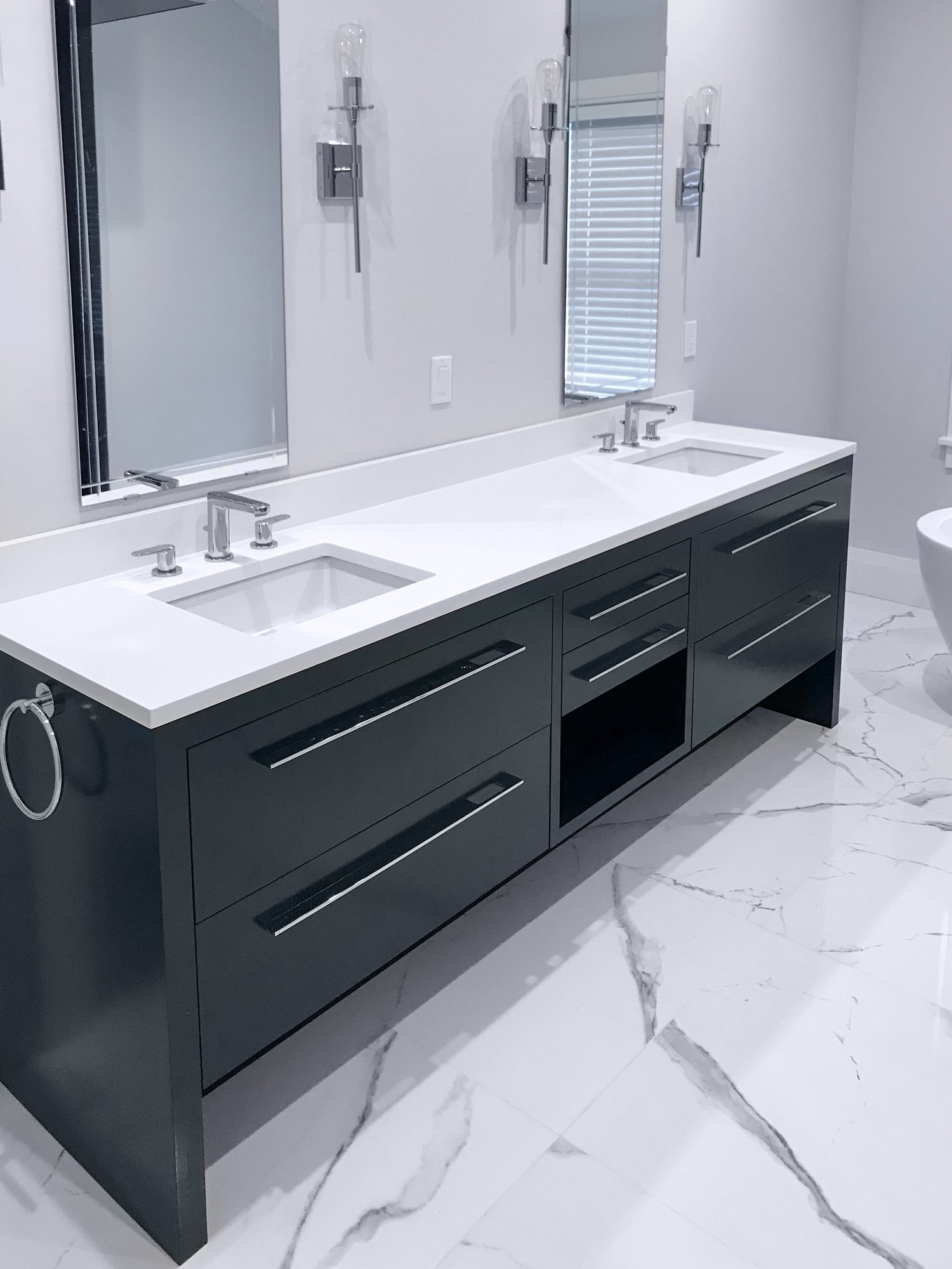 Real Modern Art Kitchen matte charcoal double vanity with slab fronts and integrated polished chrome bar pulls, sitting on a deeply recessed sculpted plinth over Calacatta-look porcelain floor, wall-mount chrome faucets, dual undermount sinks in a quartz counter